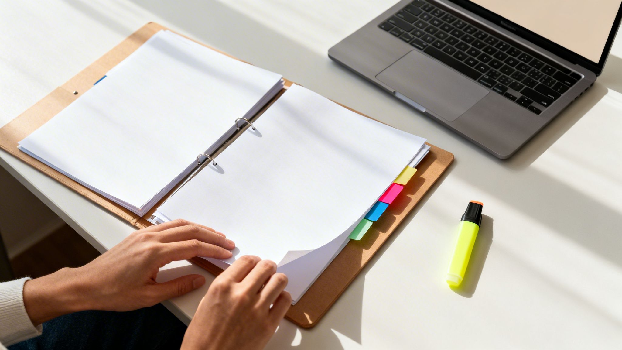 En person bladrer i et ringbind med papirer og farverige faneblade ved et skrivebord med en bærbar computer og highlighter.