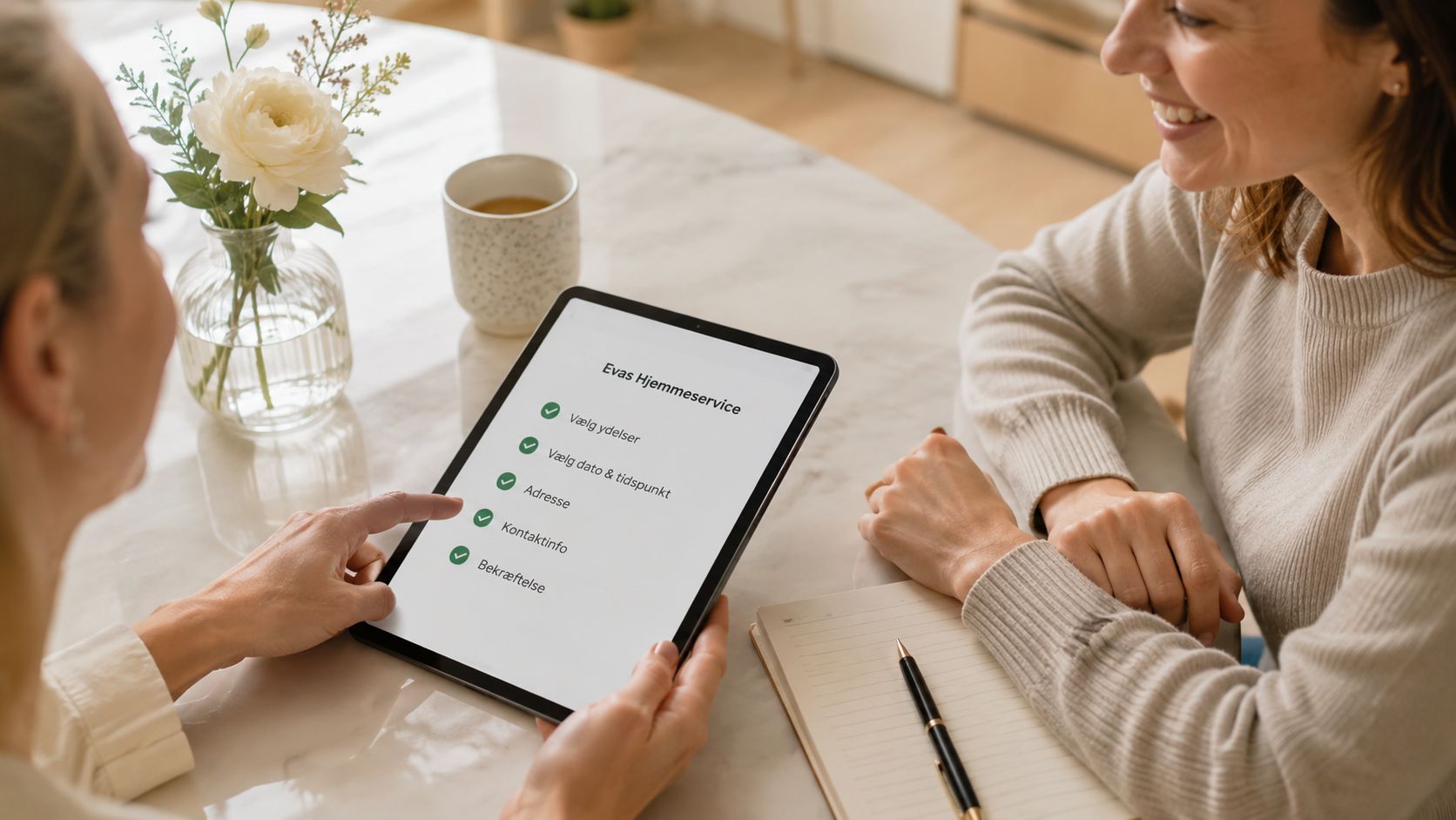 To kvinder sidder ved et bord og gennemgår en digital rengøringstjeneste på en tablet i et hjem.